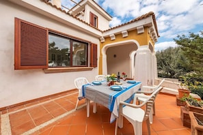 Un patio con una mesa lista para comer, rodeado de sillas blancas y una ventana con contraventanas de madera.