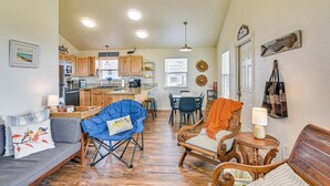 A spacious living area with a sofa, chairs, and a dining table. A kitchen with wooden cabinets and a counter is visible in the background. - Walk to Beach; Surfside Beach Home w/ Deck (Freeport)