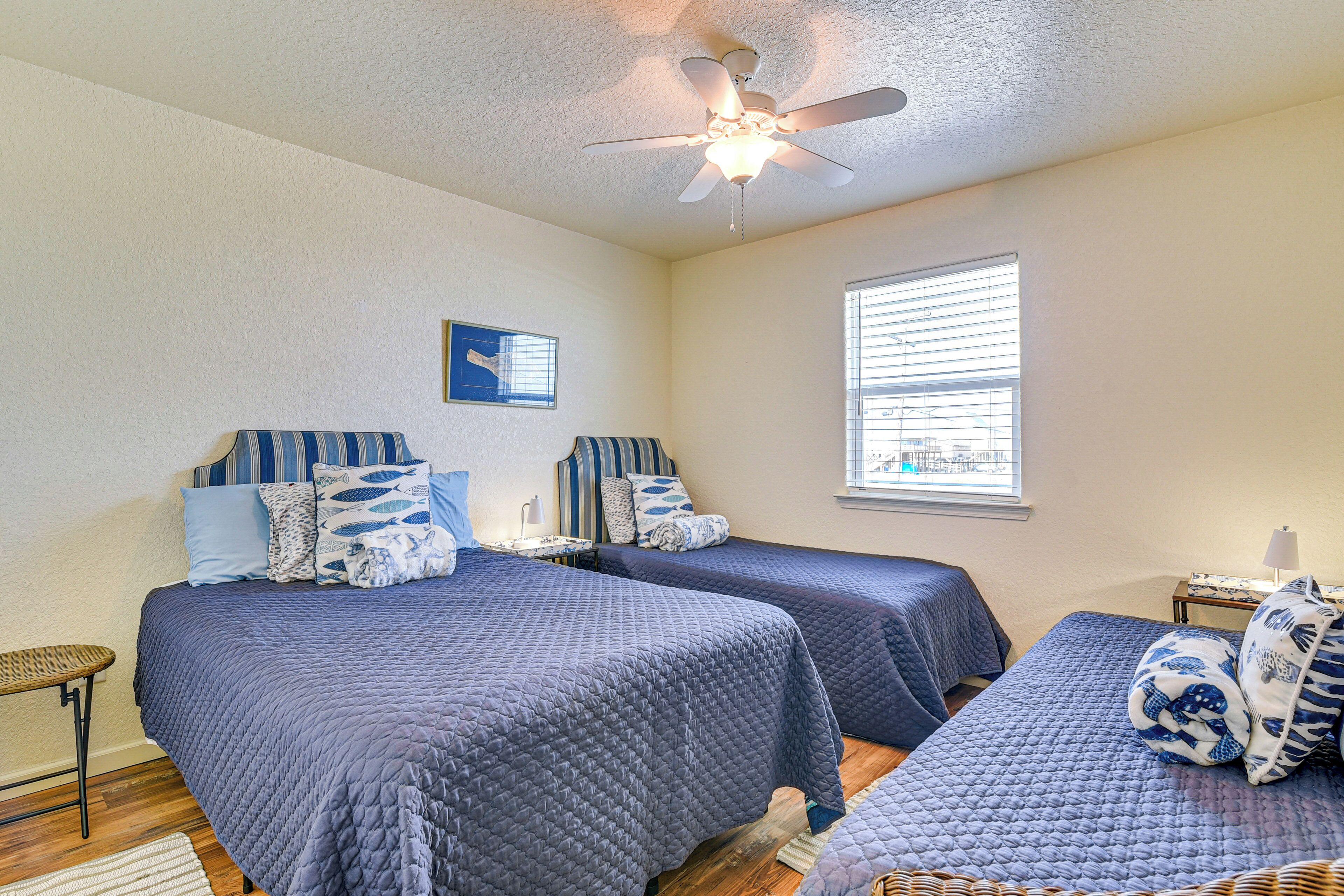 A bedroom with two beds, a ceiling fan, a window with blinds, and a small table with a lamp.