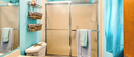 A bathroom with a glass-enclosed shower, a toilet, a sink, and a shelf with toiletries.