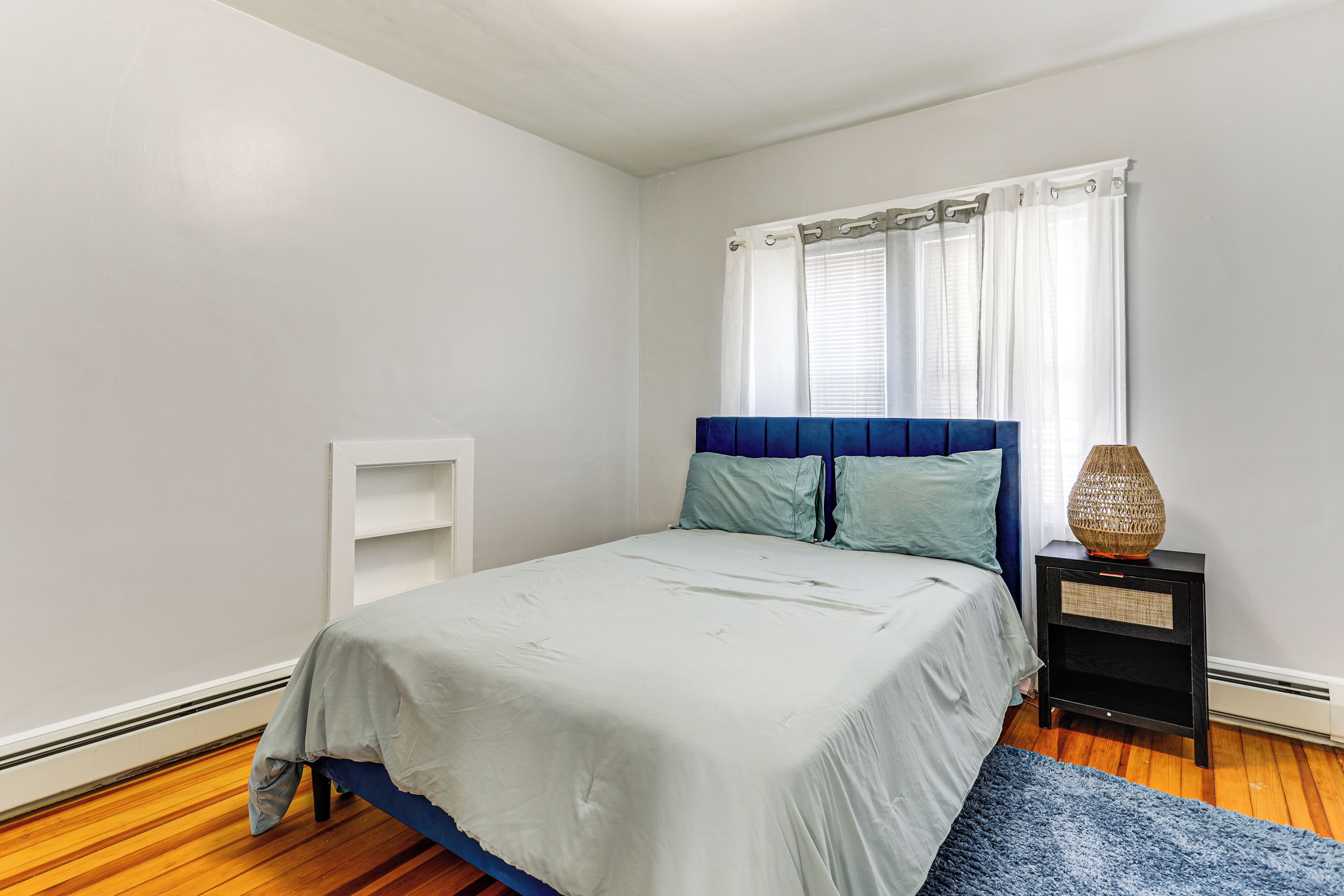A bedroom with a bed, a nightstand, a window with curtains, and a blue rug.