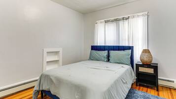A bedroom with a bed, a nightstand, a window with curtains, and a blue rug.