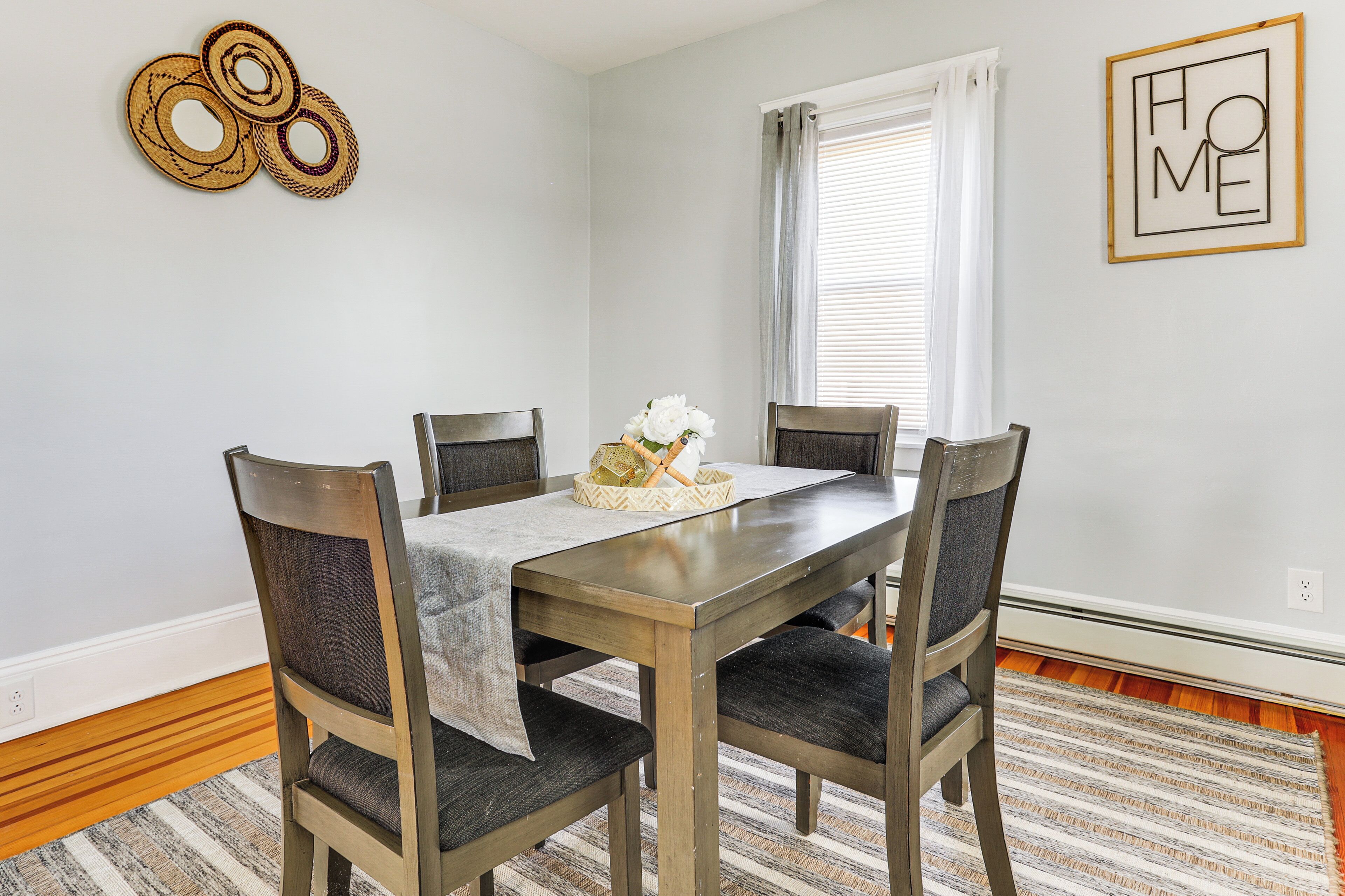 A dining area with a wooden table, four chairs, a patterned rug, a basket with flowers, and a 'HOME' sign on the wall.