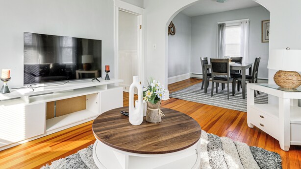Un salon comprenant un téléviseur, un meuble de rangement blanc pour appareils multimédias, une table basse ronde et un coin repas avec une table et des chaises.