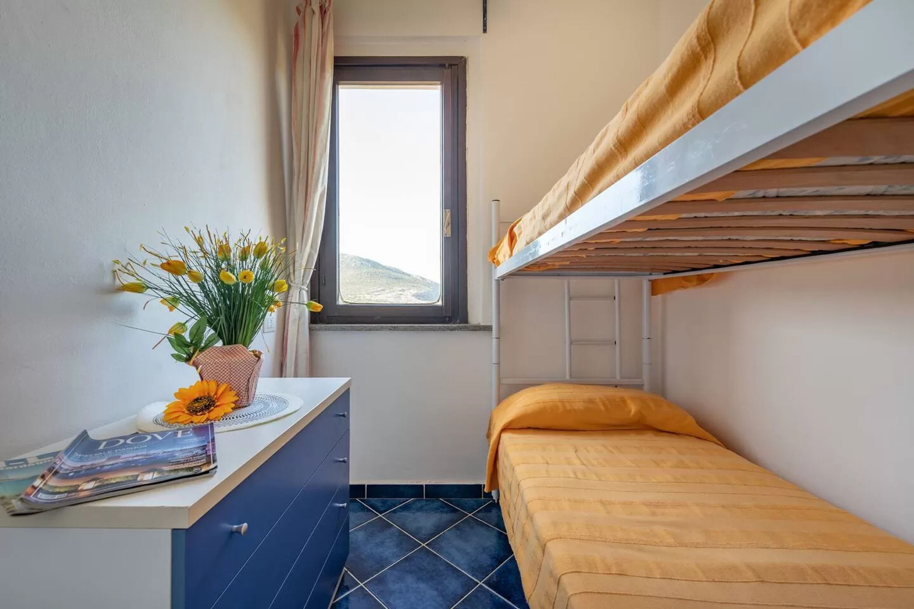 A small, compact room with a bunk bed, a desk with a vase of flowers, and a window providing a view of the outdoors.