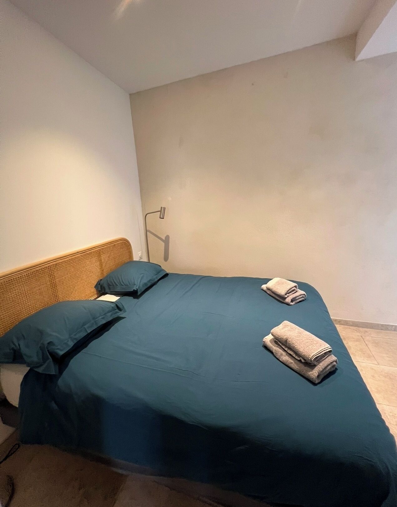 A small, single-bed room with a woven headboard, a bedside lamp, and neatly folded towels on the bed.