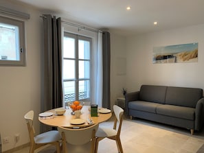 Ein Essbereich mit einem runden Tisch, Stühlen und einem Couchtisch. Im Hintergrund steht eine Couch und ein Fenster mit Vorhängen.