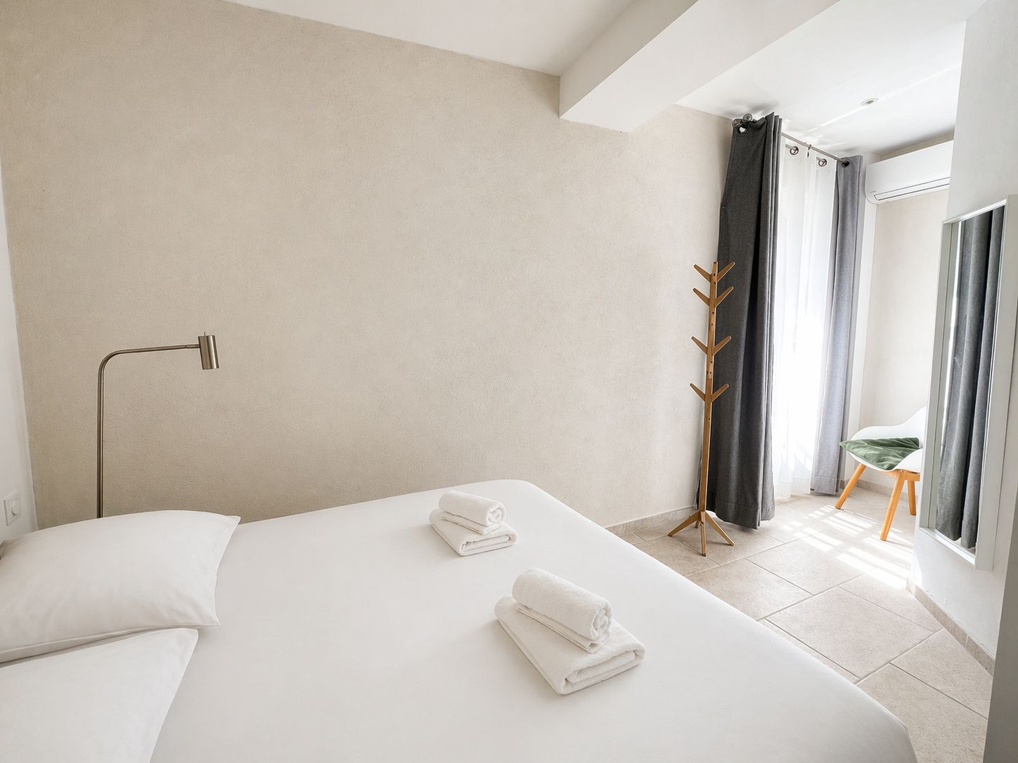 Hotellihuone, jossa on sänky, tuoli, naulakko ja ikkuna, jossa on verhot.
