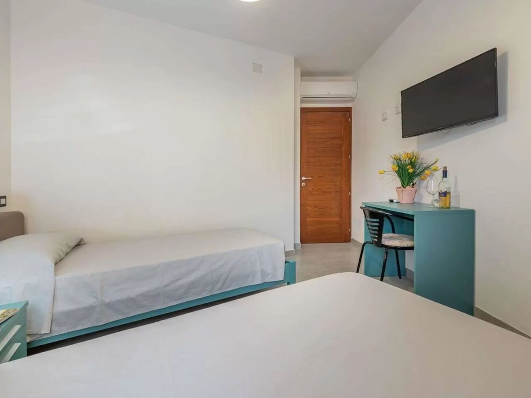 A room with two beds, a desk with a chair, a flat-screen TV, and a wooden door.