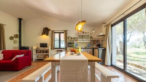 Un salon chaleureux avec un canapé rouge, une table à manger en bois avec des bancs, un coin cuisine doté d’une grande fenêtre et une cheminée.