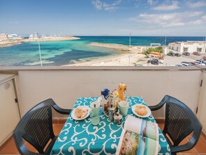 Varanda com mesa posta para o café da manhã, com vista para uma praia e um porto.
