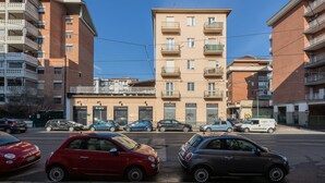 Un edificio residencial de varios pisos con balcones, una zona comercial en la planta baja con tiendas, y coches estacionados en la calle.