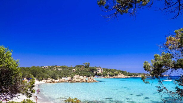 Una playa costera con agua turquesa cristalina, costa rocosa y edificios en una colina.