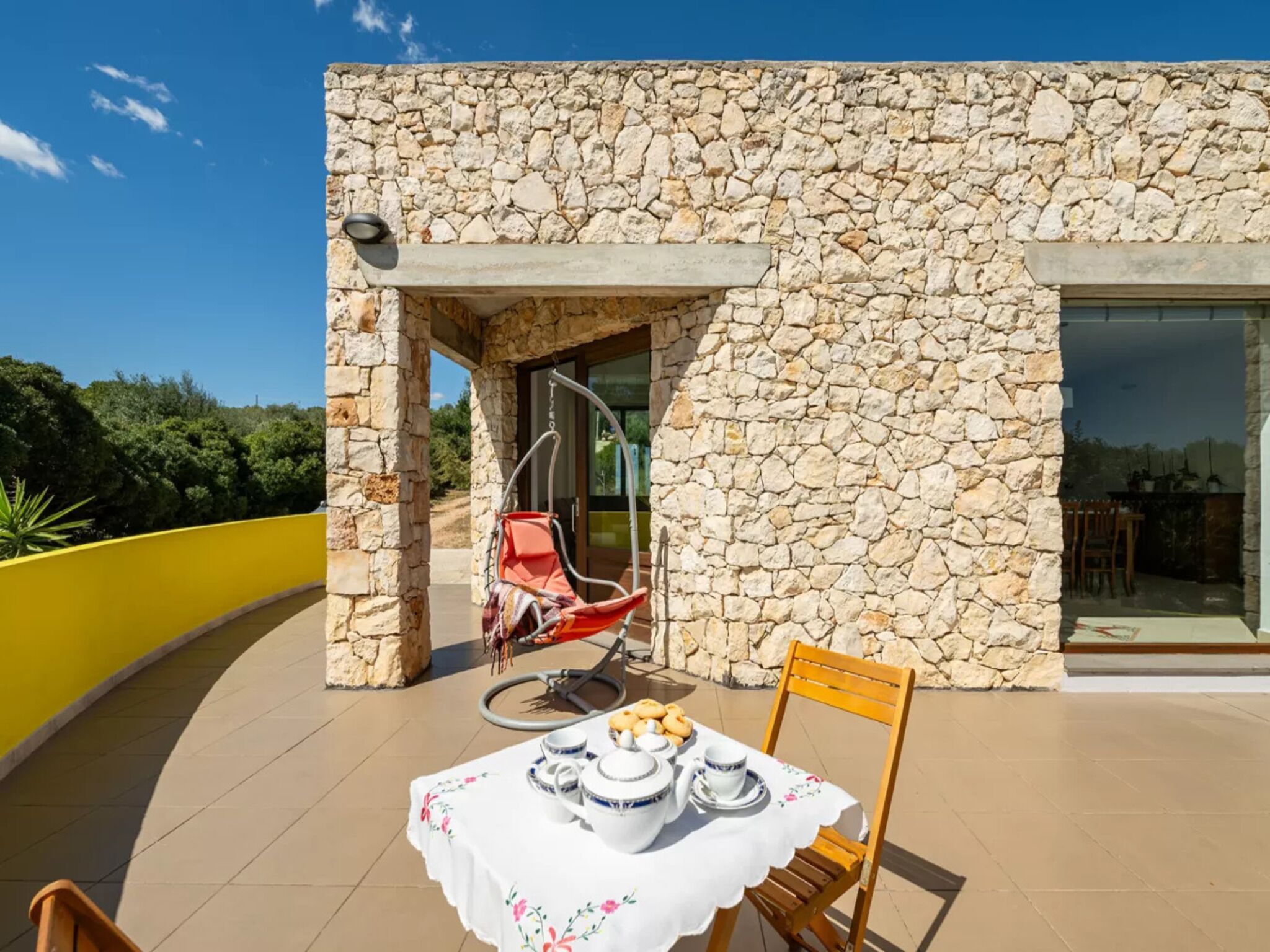 A stone building with a patio area featuring a table set for tea, a hanging chair, and a yellow wall.