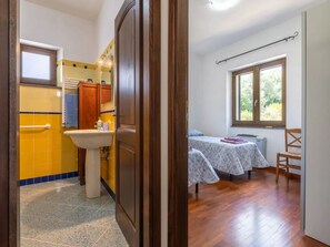 Een badkamer met gele tegels, een wastafel en een spiegel. Een houten deur leidt naar een slaapkamer met een bed en een raam.