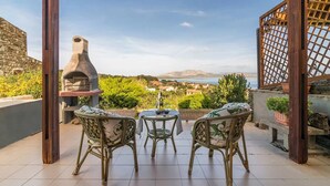 Un área de estar al aire libre techada con sillas de mimbre y una mesa, con vistas a un paisaje pintoresco que incluye el mar y montañas distantes.