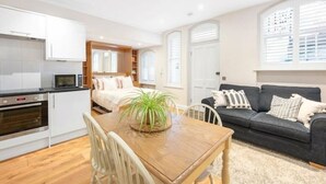Een ruimtelijk ingerichte leefruimte met kitchenette, eethoek en zithoek met bank en potplant.