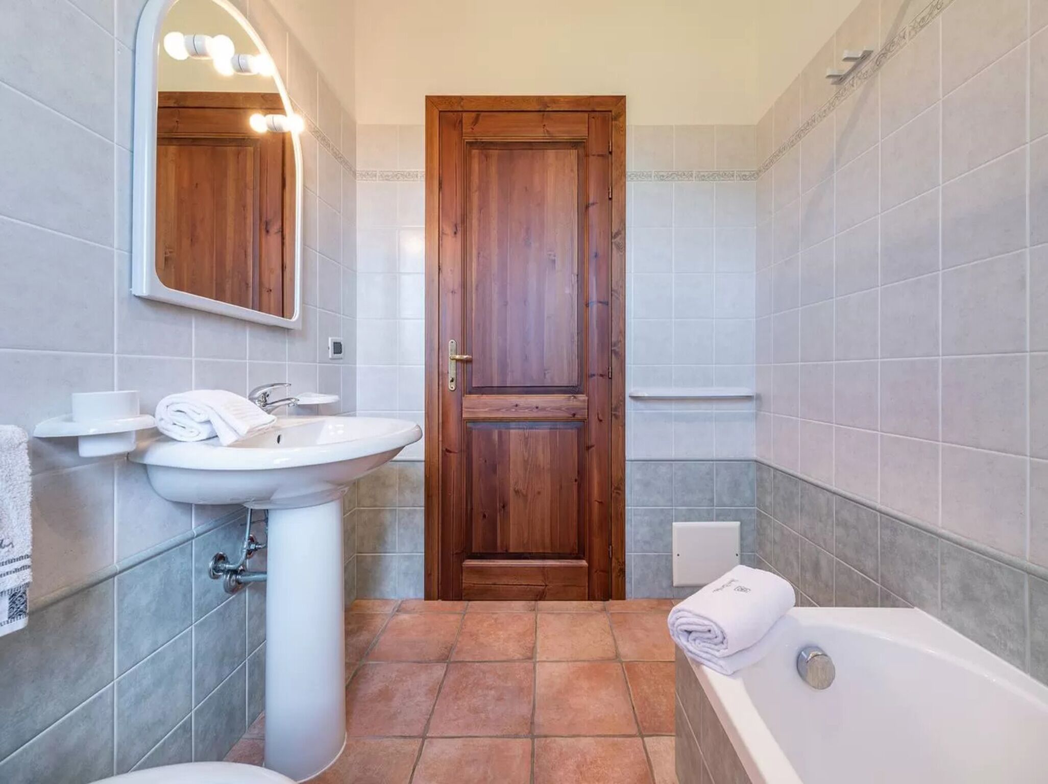 Un baño con puerta de madera, un lavabo blanco, un espejo con luces y una bañera.