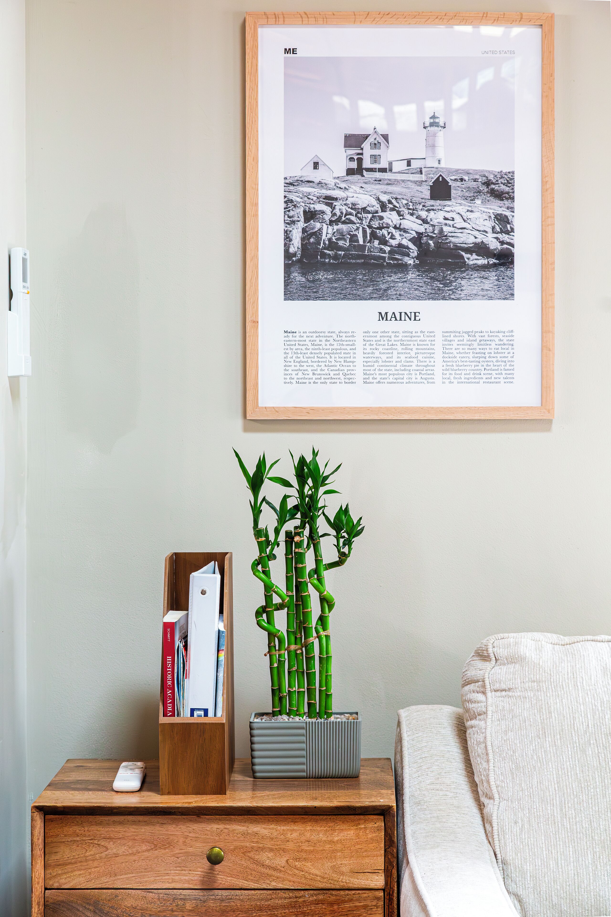 Fotografia em preto e branco emoldurada de um farol e edifícios em Maine, uma mesa de cabeceira de madeira com um controle remoto, uma planta de bambu e um livro.
