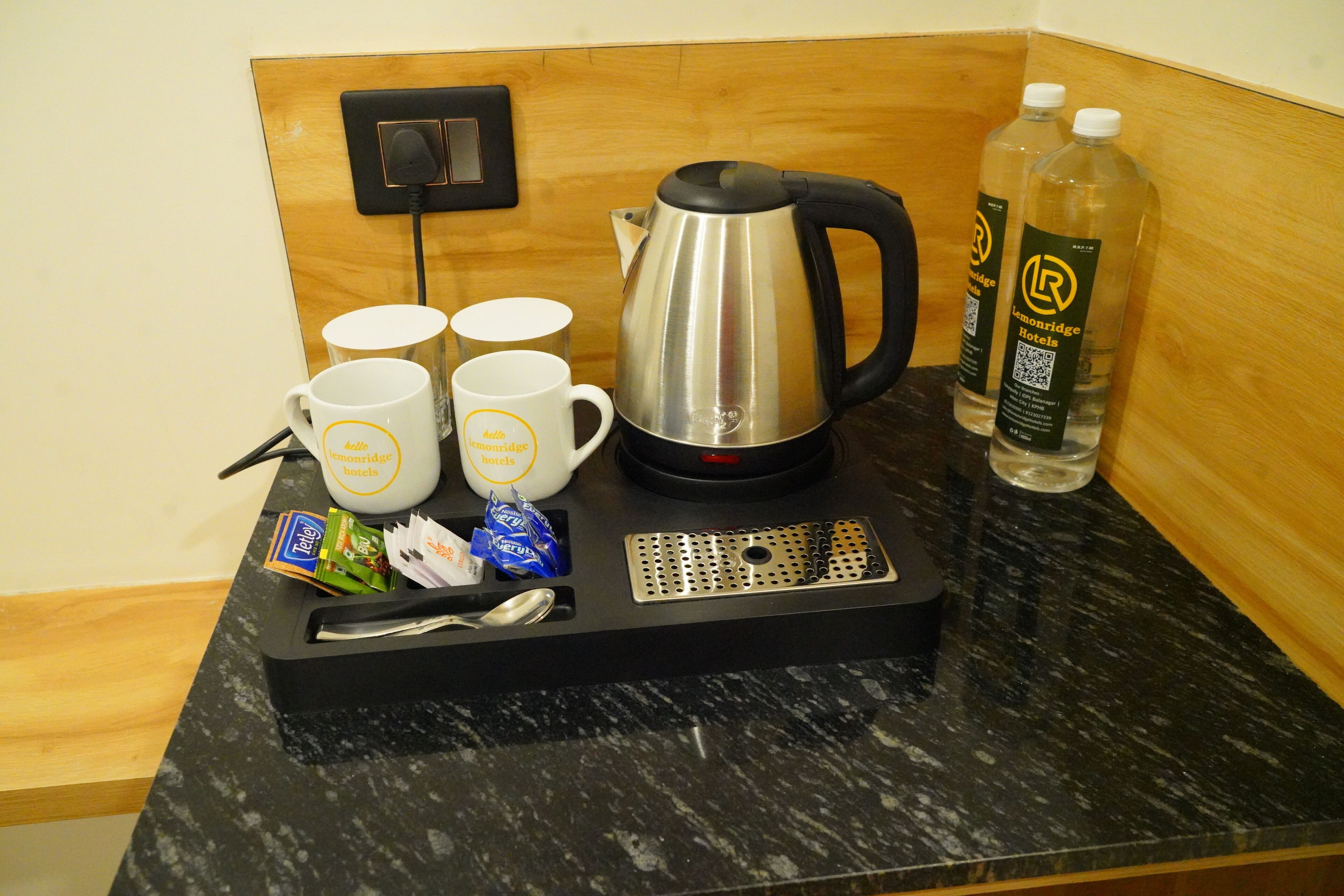 A coffee station with a kettle, four mugs, a tray with condiments, and two bottles of water.