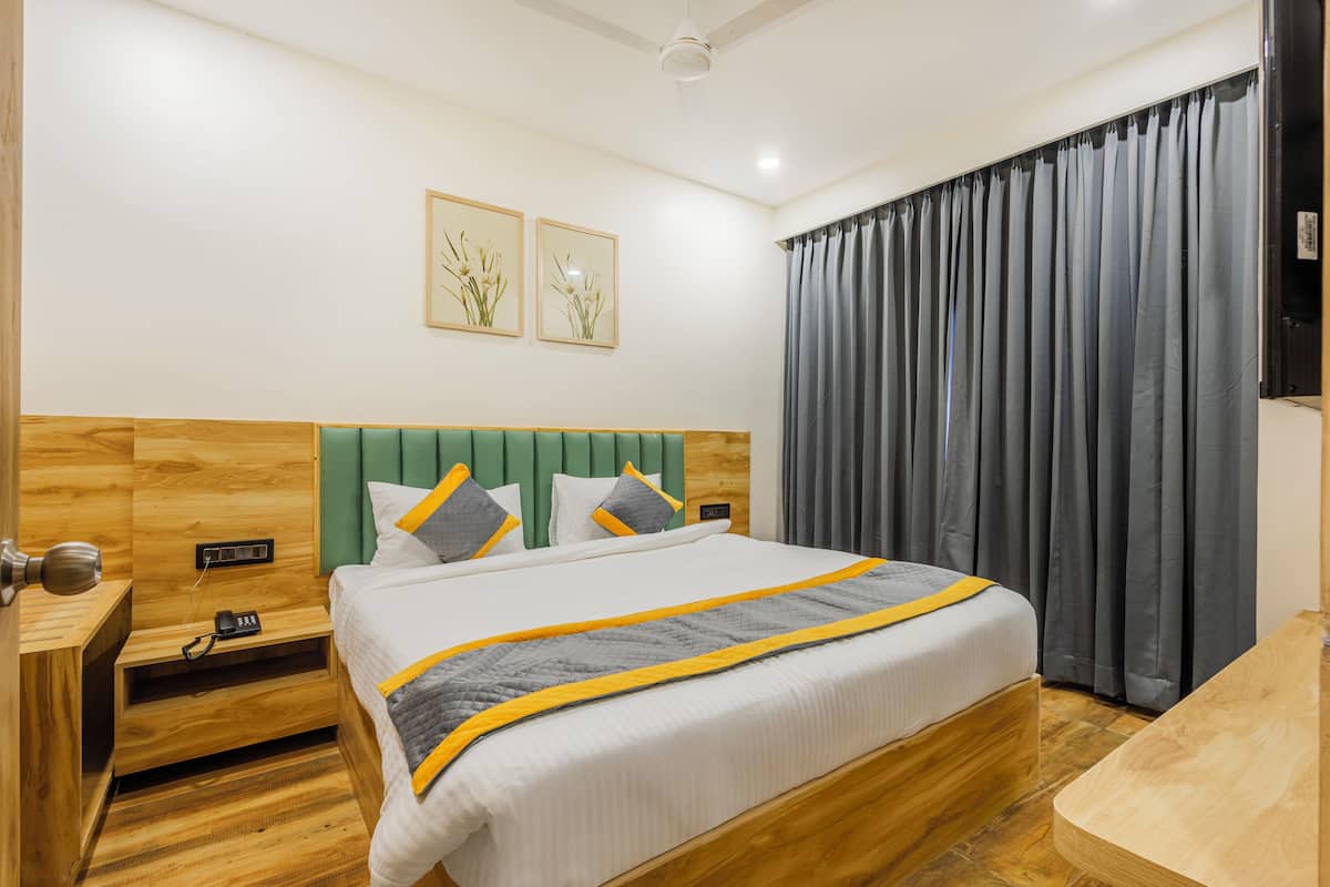 Habitación de hotel con cabecera de madera, cama con colcha gris y amarilla, dos cuadros con flores enmarcadas en la pared y una cortina gris en la ventana.