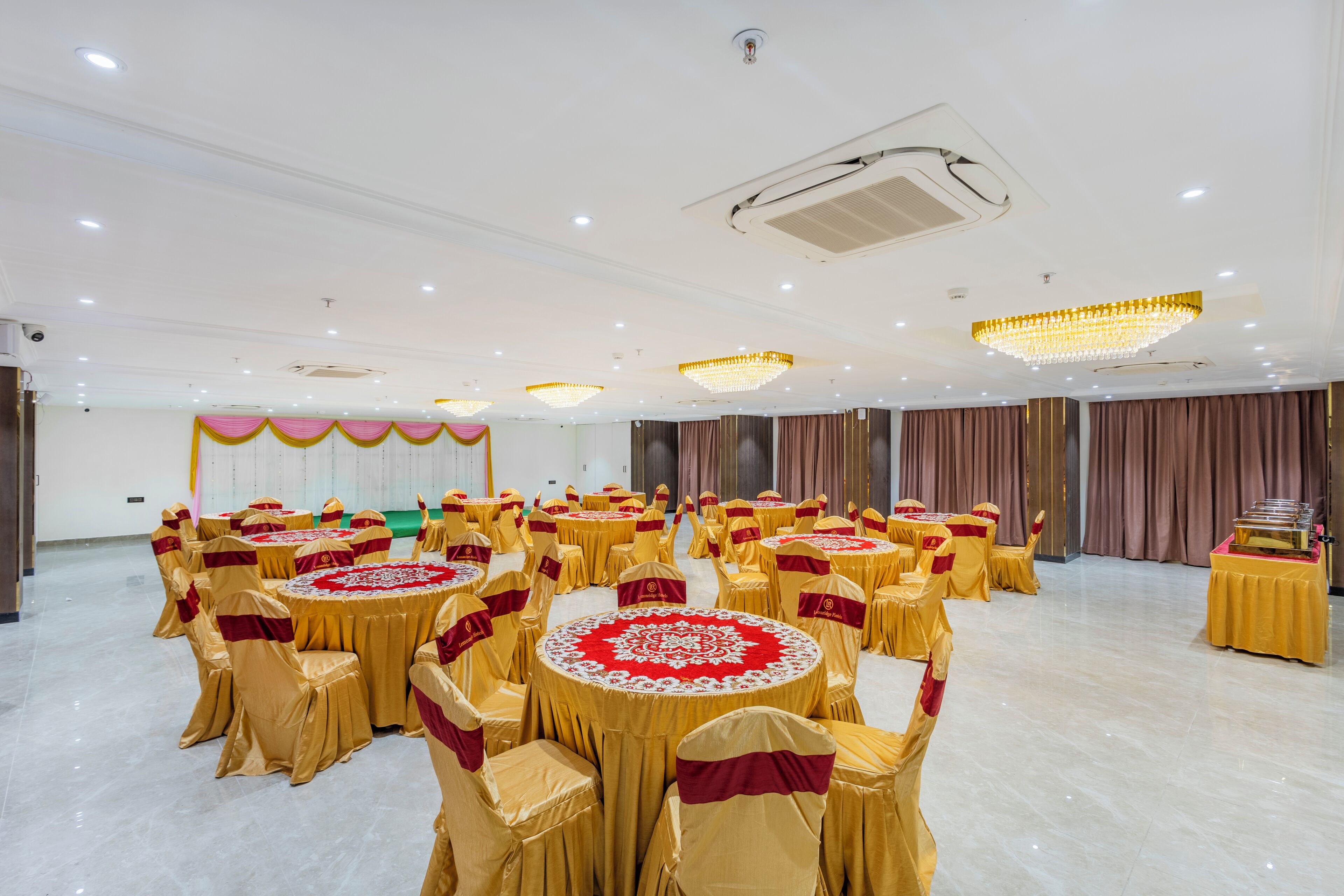 Un salón de banquetes con mesas redondas cubiertas con manteles dorados y centros de mesa rojos, dispuestas en filas.