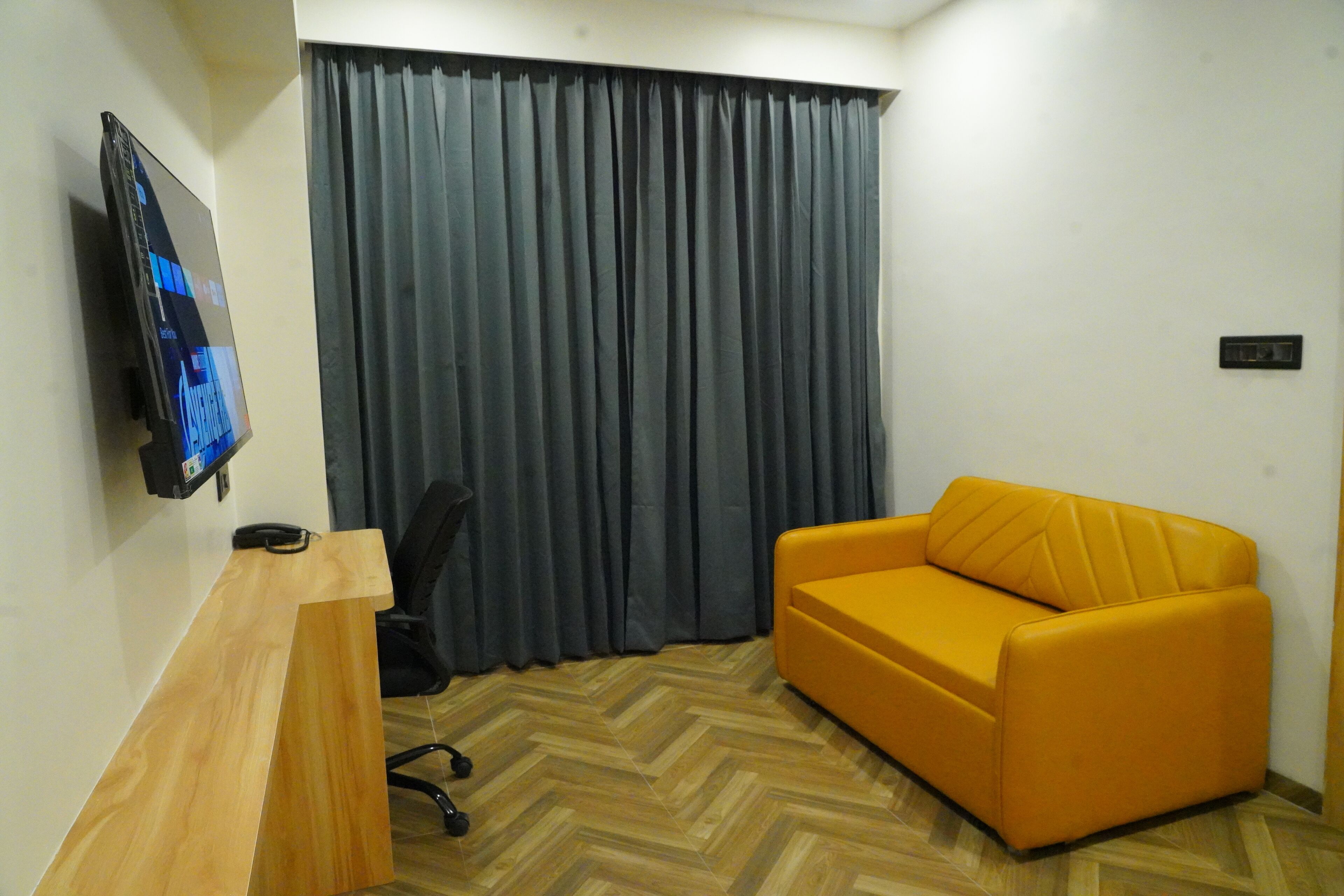 A room with a wooden desk, a black office chair, a yellow sofa, a television on the wall, and a large grey curtain.