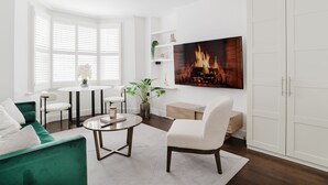 A modern living room with a green sofa, a white armchair, a round coffee table, and a fireplace with a fire. - Spacious Luxury Flat with Garden in Central London (London)