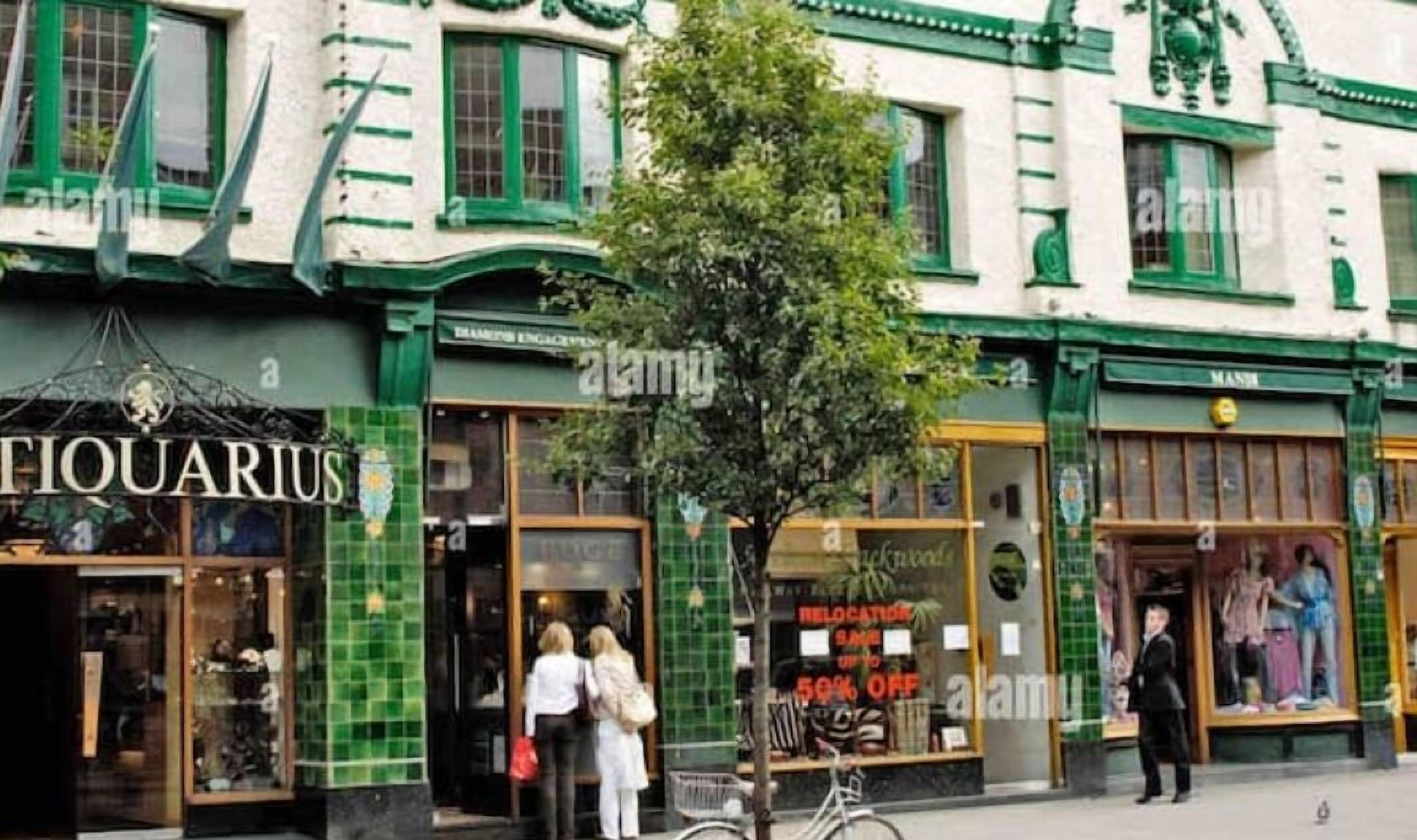 A building with a green and white facade, storefronts with large windows, and a tree in front.