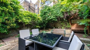 A patio with wicker furniture and a glass table, surrounded by lush greenery and a brick wall. - Spacious Luxury Flat with Garden in Central London (London)