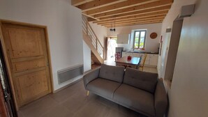 A spacious living area with a sofa, wooden ceiling beams, a staircase, and a kitchen in the background.