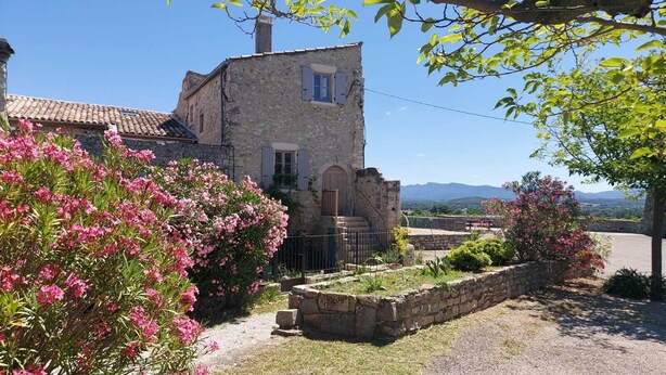 A stone building with a tiled roof, surrounded by flowering bushes and a stone wall.