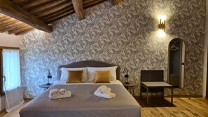 A hotel room with a patterned wallpaper, a bed with pillows, a bedside table with a lamp, and a small table with towels. - Il Bivio (Ostellato)
