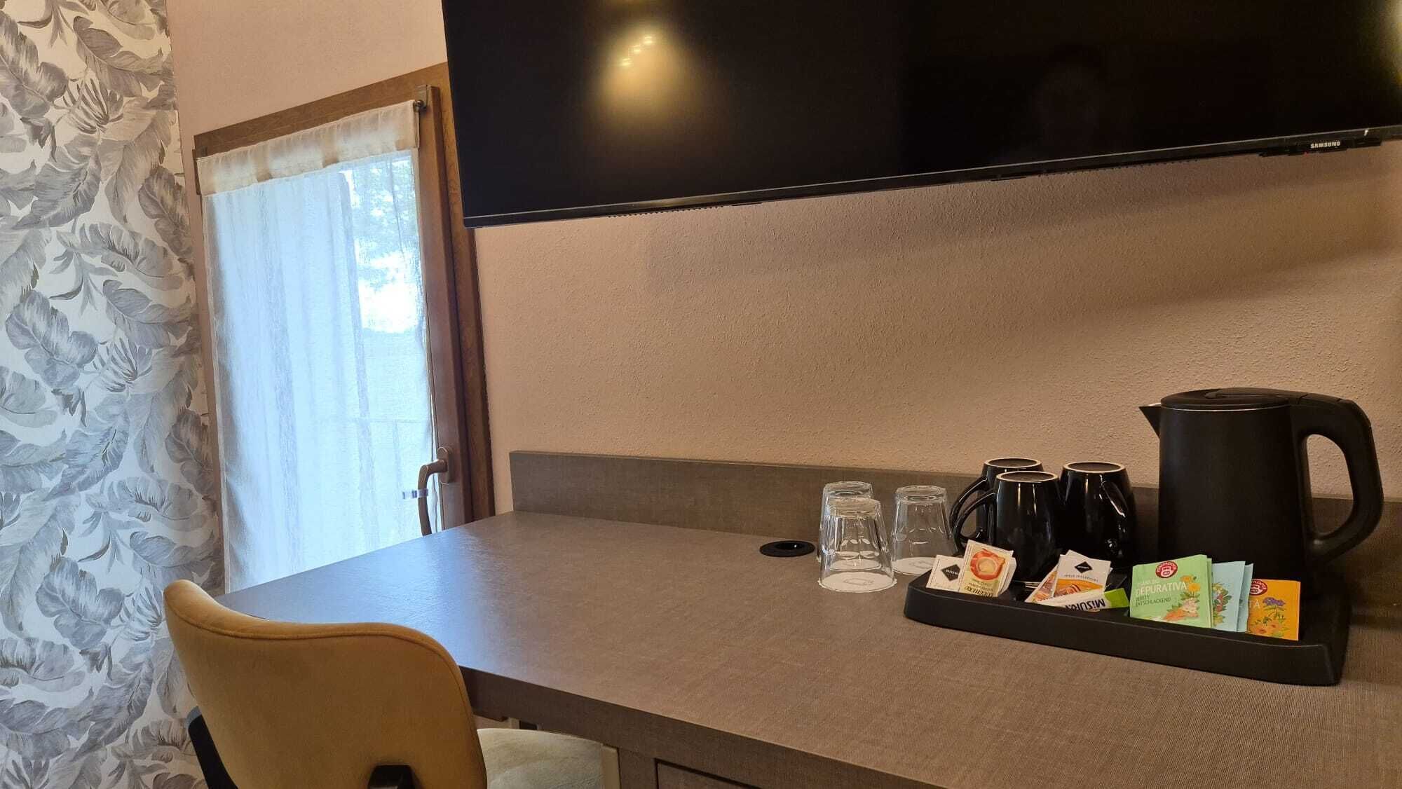 Ein Hotelzimmer mit Flachbildfernseher an der Wand, einem Tisch mit einer Kaffeestation und einem Fenster mit Vorhang.