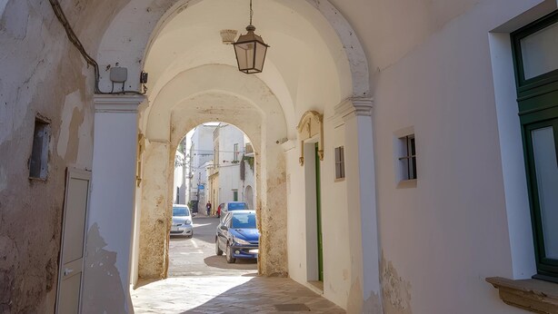 Un corredor abovedado con un farol colgante, que conduce a una calle con coches estacionados y edificios al fondo.