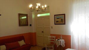 Una habitación con un sofá rojo, una mesa blanca, una araña y una puerta, además de dos cuadros en las paredes.
