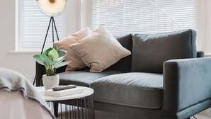 A modern living room with a grey sofa, two cushions, a small round table with a plant and a book, and a floor lamp. - Luxury Flat in a Great Location / Harrods (London)