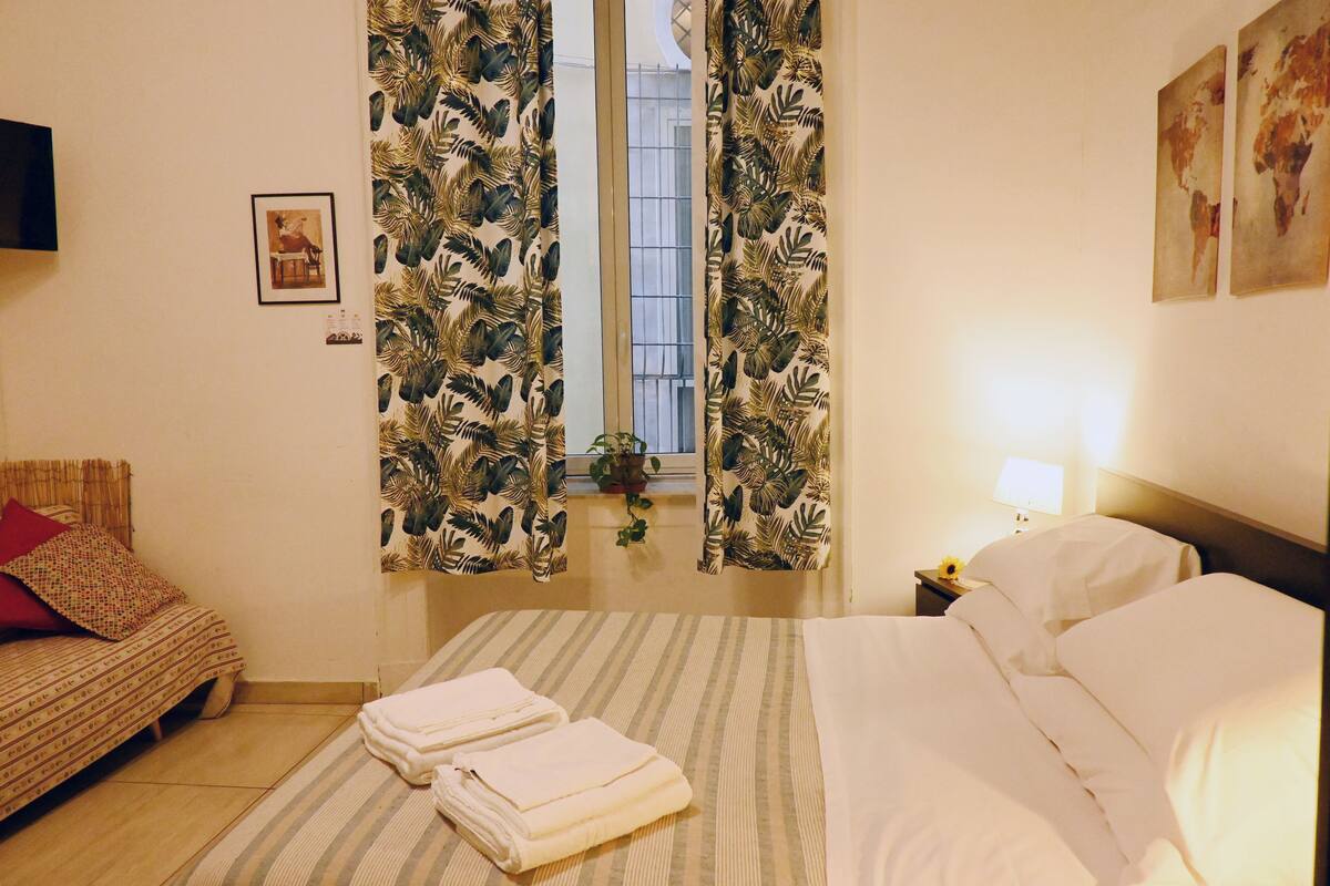 Ein Hotelzimmer mit einem Bett, einem kleinen Tisch mit Lampe, einem Fenster mit Vorhängen und einem an der Wand befestigten Fernseher.