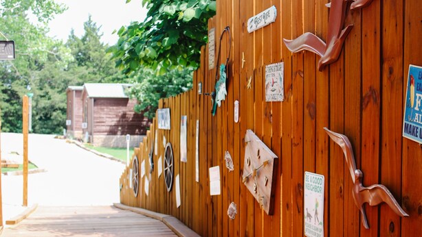 A wooden fence with various signs and decorations, including a 'Be a Good Neighbor' sign and a bird sculpture.