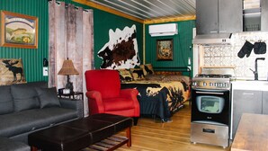 A cozy living room with a sofa, a red armchair, a wooden coffee table, a patterned bedspread, and a kitchen area with a stove and oven.