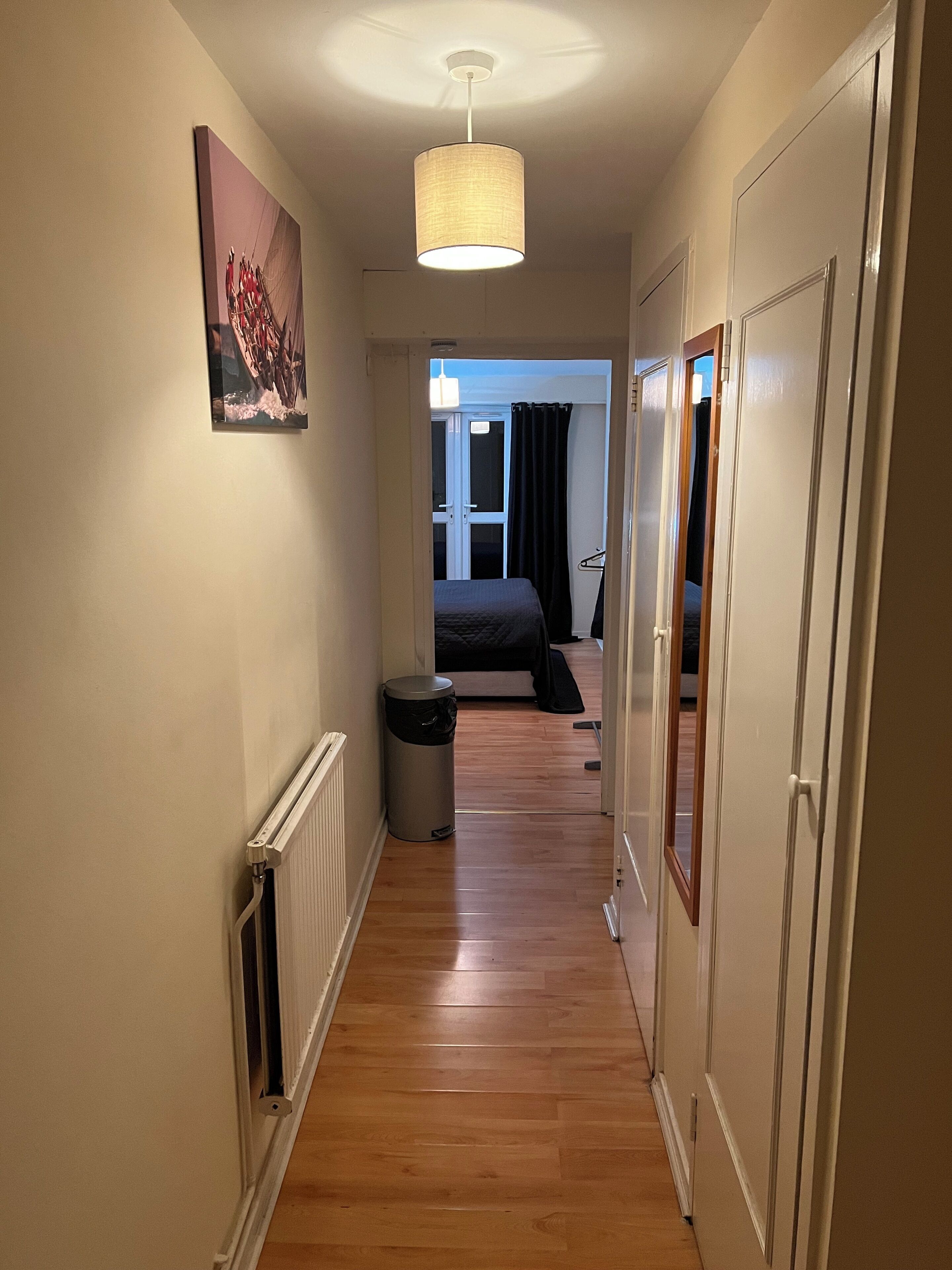 Un pasillo estrecho con suelo de madera, un radiador, un cesto de basura y una cama en la habitación contigua.