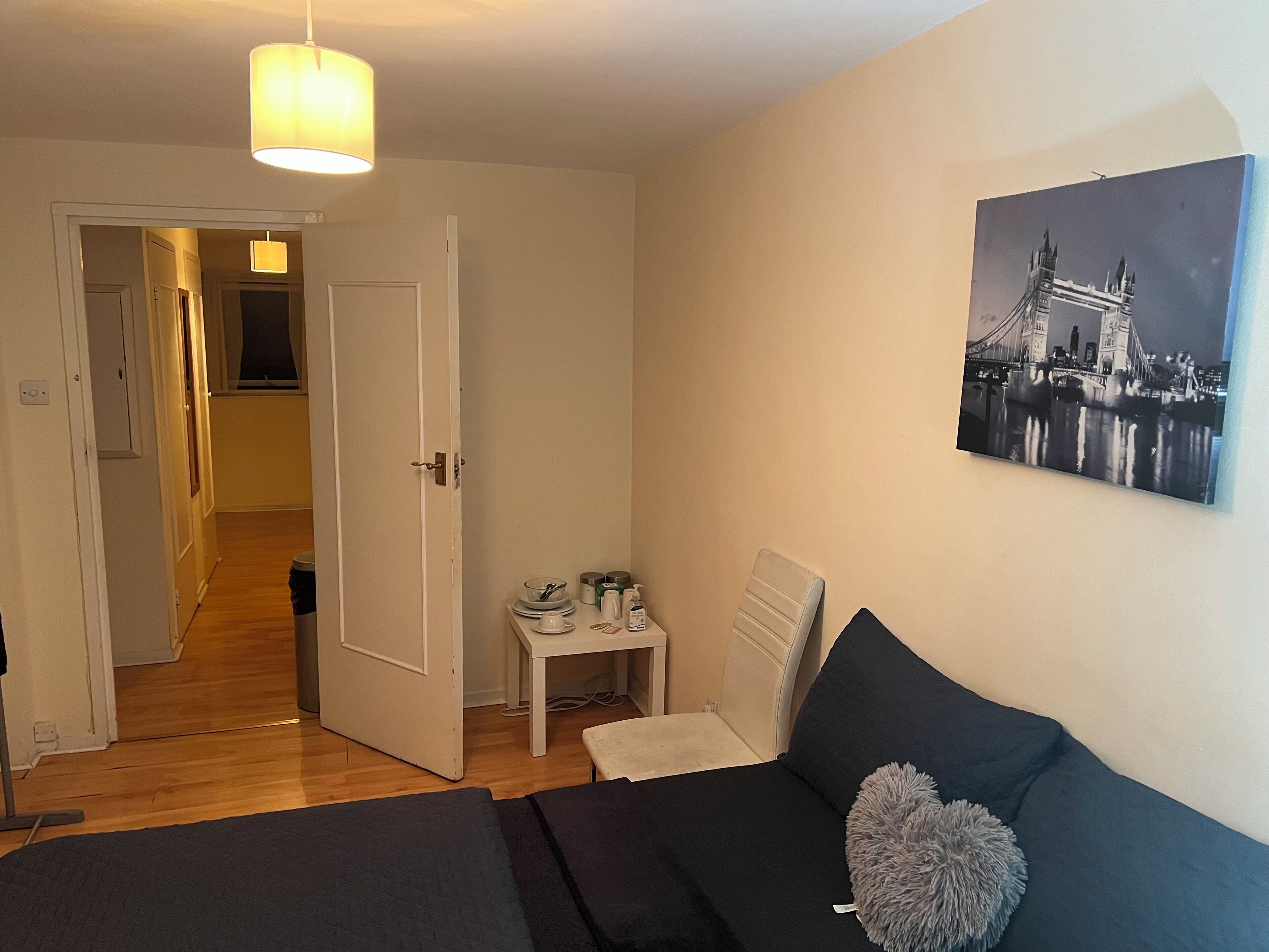 Une chambre à coucher comprenant un lit, une table de chevet, une lampe, un tableau représentant un pont et une porte donnant sur une autre pièce.