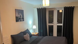 A bedroom with a bed, a nightstand, a lamp, a window with curtains, a chair, and a painting on the wall. - 1 double room in London with WiFi (London)