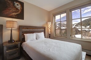 Un dormitorio con una cama grande, cabecera de madera, mesita de noche con lámpara y vistas a un paisaje montañoso nevado a través de un ventanal amplio.
