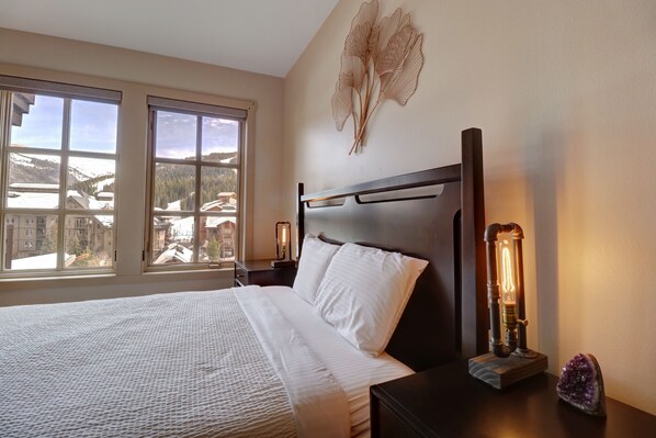 Un lit bien fait, avec du linge de lit blanc, une tête de lit sombre et une table de chevet sur laquelle se trouvent une lampe et un objet décoratif en cristal.