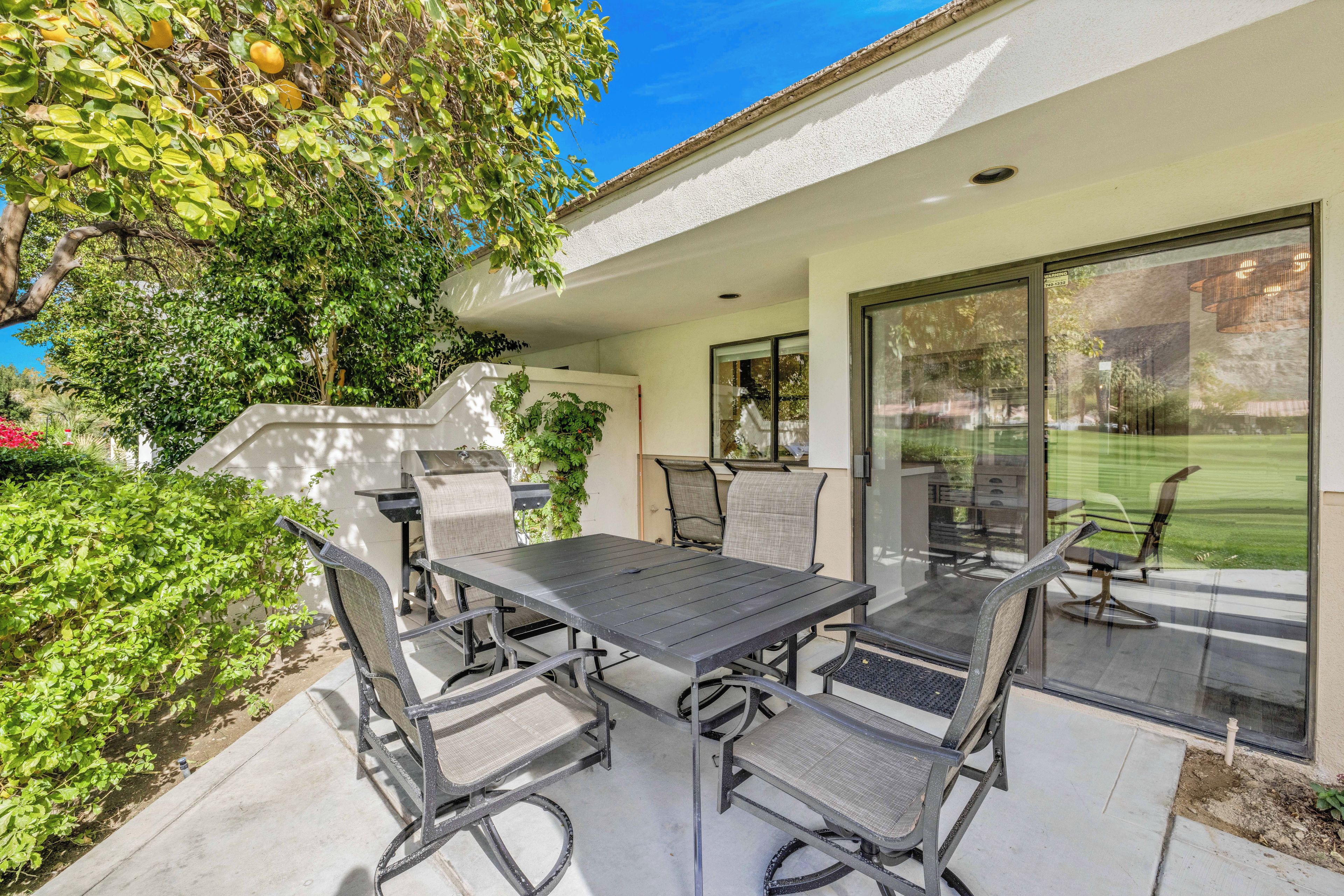 An outdoor patio with a table and chairs, surrounded by greenery and a view of a lawn.