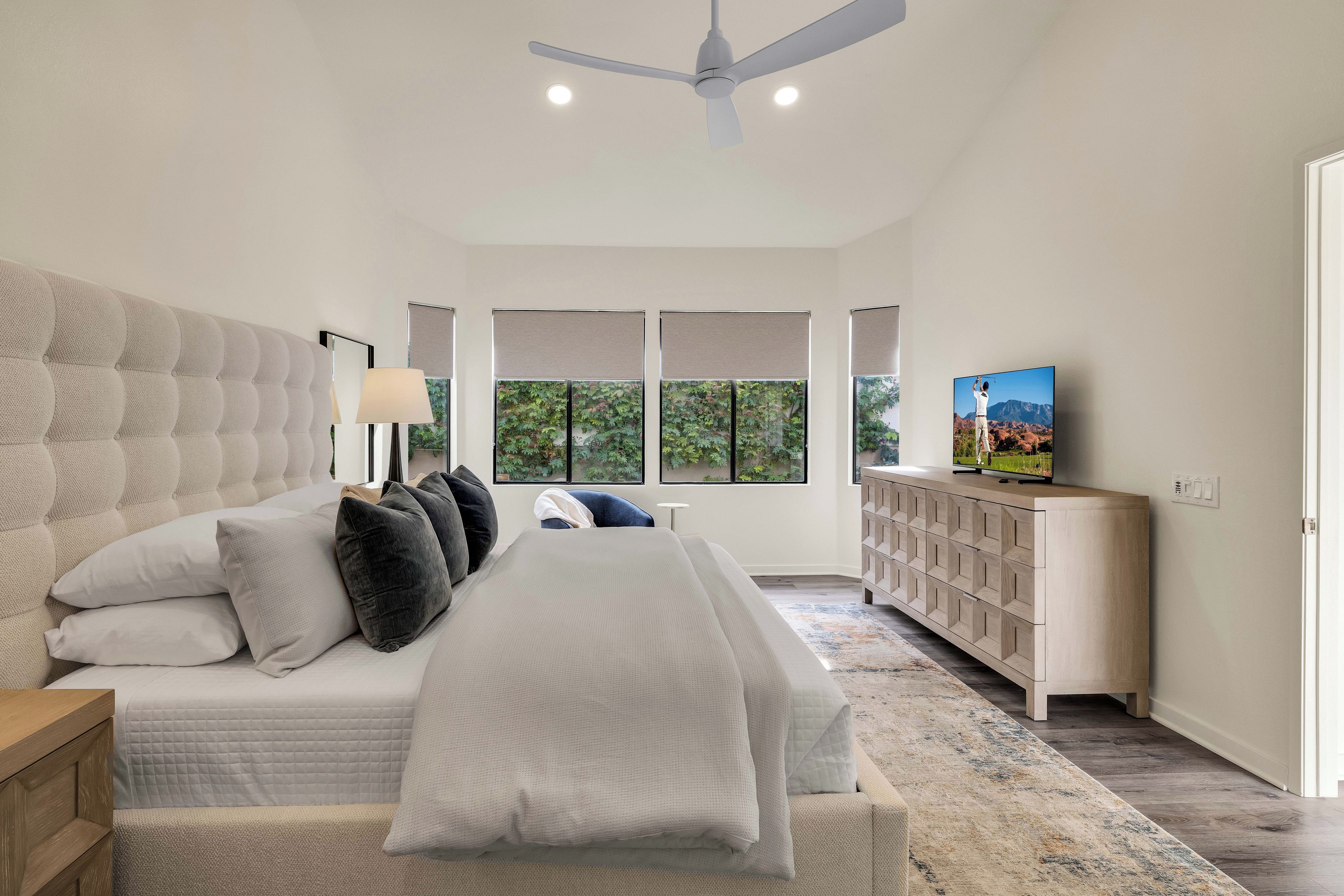 Une chambre avec un grand lit, un ventilateur de plafond, un téléviseur posé sur un meuble en bois, et une vue sur la verdure par les fenêtres.