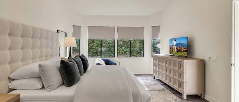 A bedroom with a large bed, a ceiling fan, a TV on a wooden cabinet, and a view of greenery through the windows.