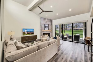 Ein modernes Wohnzimmer mit einer großen Eckcouch, einem Flachbildfernseher, einem Kamin und Blick auf einen Golfplatz durch bodengrosse Fenster.