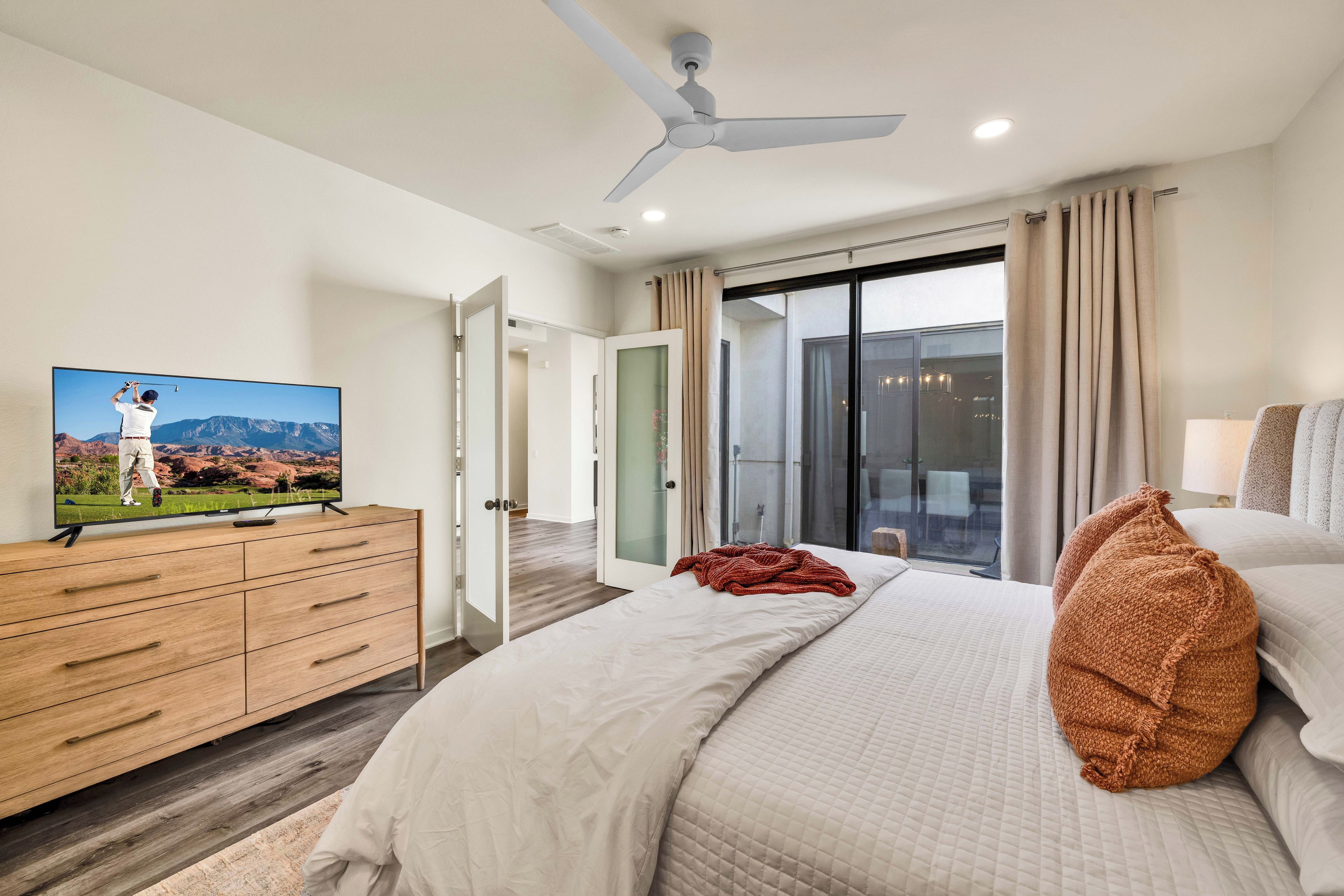 Une chambre d’hôtel moderne équipée d’un lit, d’un téléviseur posé sur une commode en bois, d’un ventilateur de plafond et offrant une vue sur l’extérieur grâce à une porte coulissante.